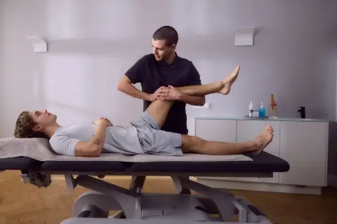 Physiotherapeut dehnt das Bein eines Patienten auf einer Liege. Die Knie-Schiene nach OP wird sanft bewegt, um die Beweglichkeit wiederherzustellen.