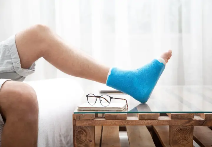 Mann mit blauem Gipsbein ruht sich aus. Die Frage "welche schuhe nach fersenbeinbruch" beschäftigt ihn, während er auf einem Holztisch mit Glasplatte entspannt.