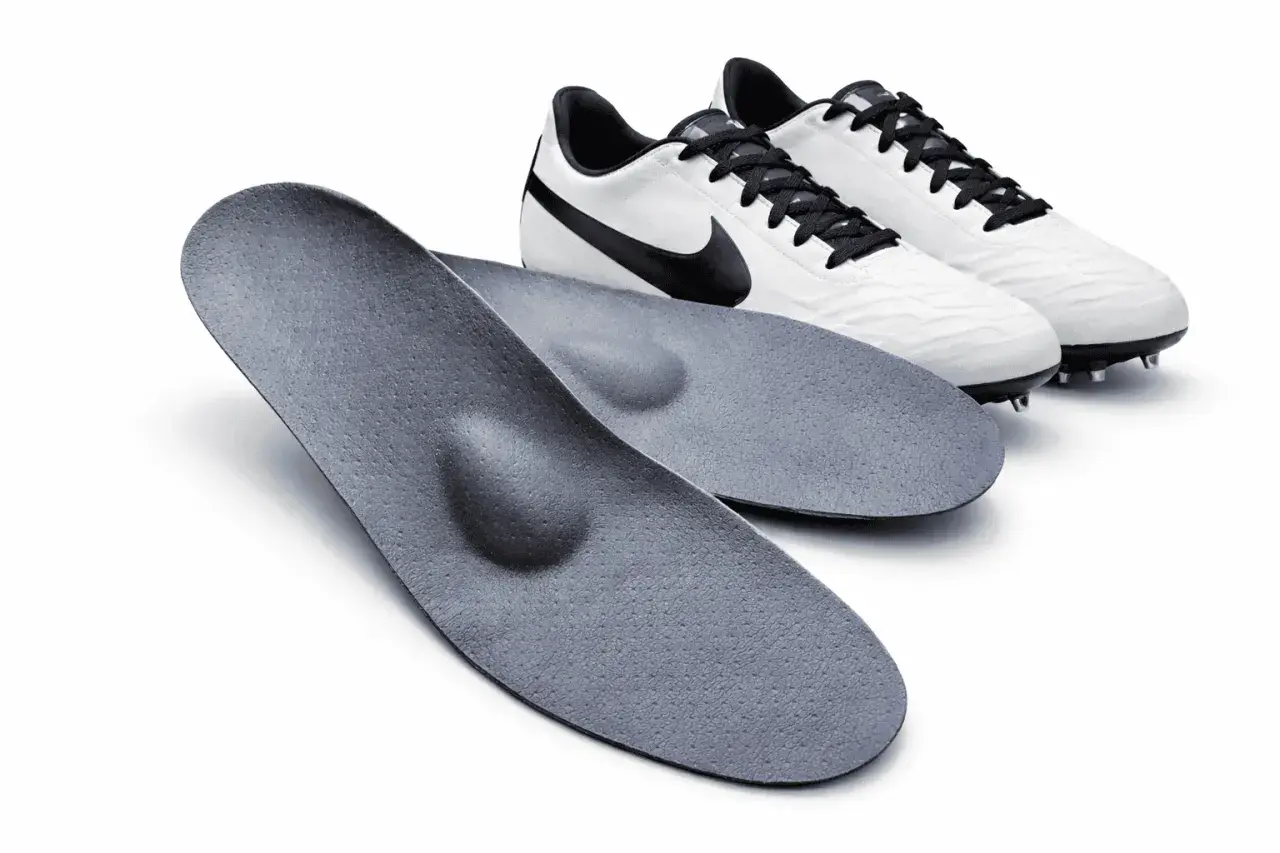 Zwei orthopädische fußball einlagen liegen vor einem Paar weißer Fußballschuhe mit schwarzen Schnürsenkeln und Swoosh-Logo.