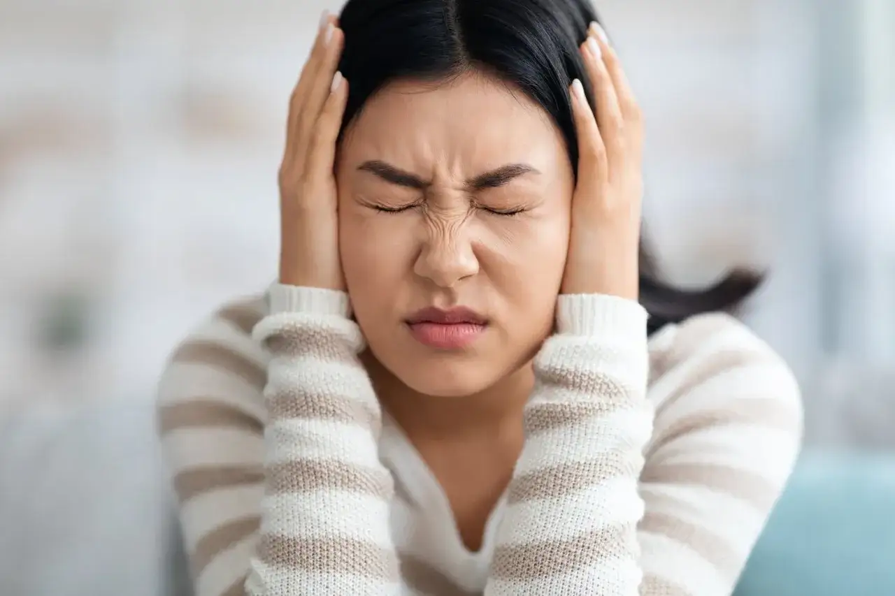 Frau mit geschlossenen Augen und H&auml;nden an den Ohren, die unter Tinnitus leidet. Osteopathie kann helfen.