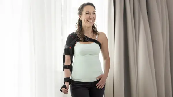 Lächelnde Frau mit einer Schulterbandage, die ihr linkes Schultergelenk fixiert.